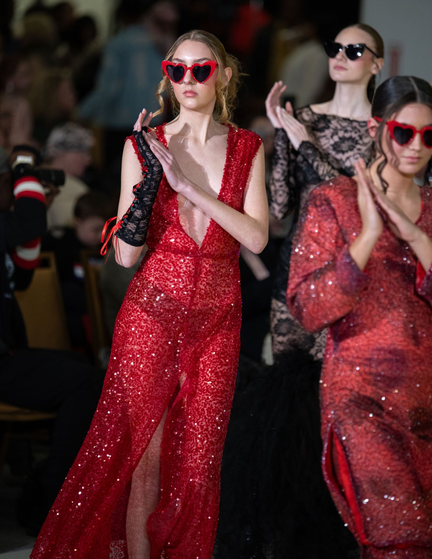 Sparkling red gown and pink sunglasses displayed on runway with audience in background.