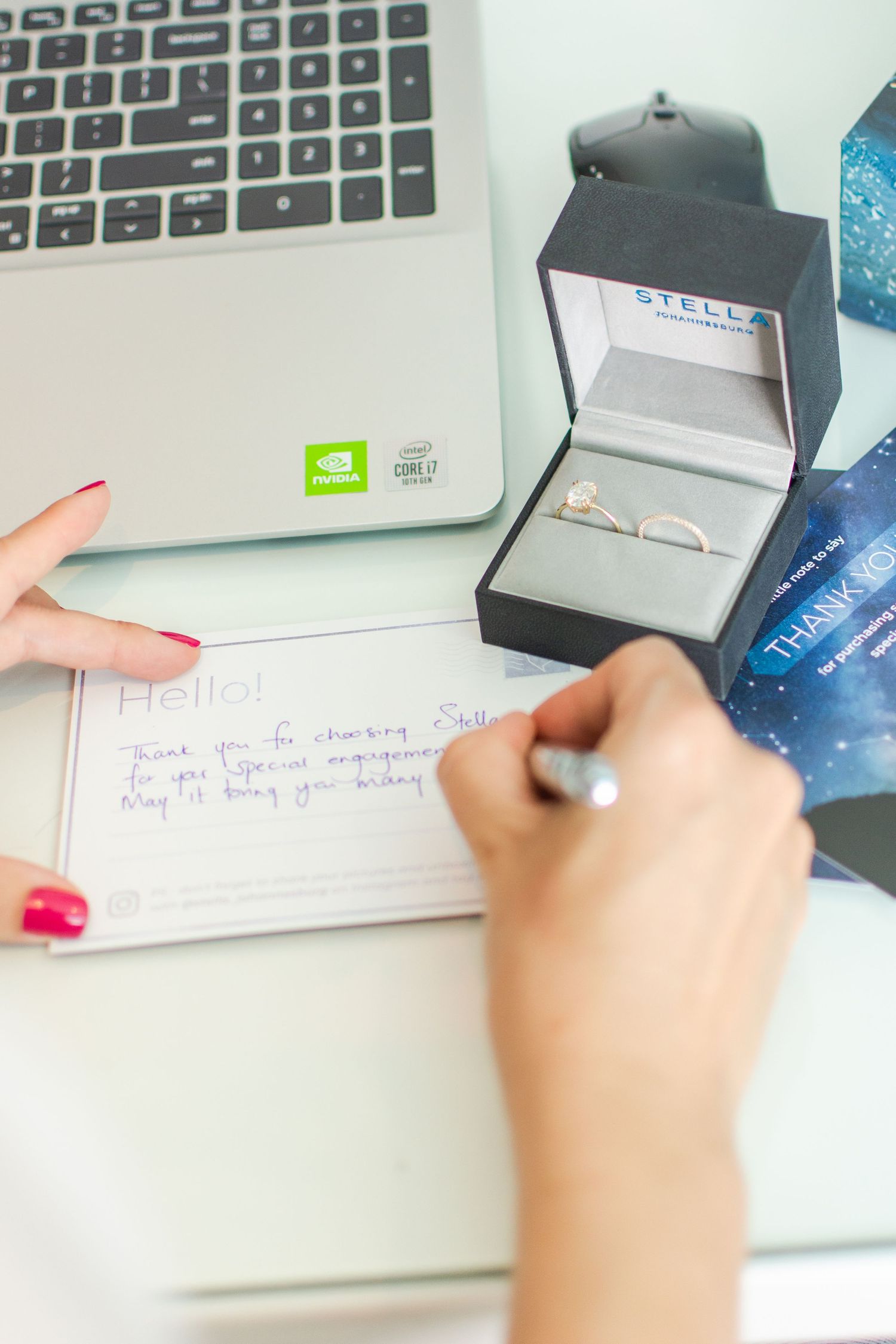 Close up of hands writing note next to laptop and jewelry box on white desk.