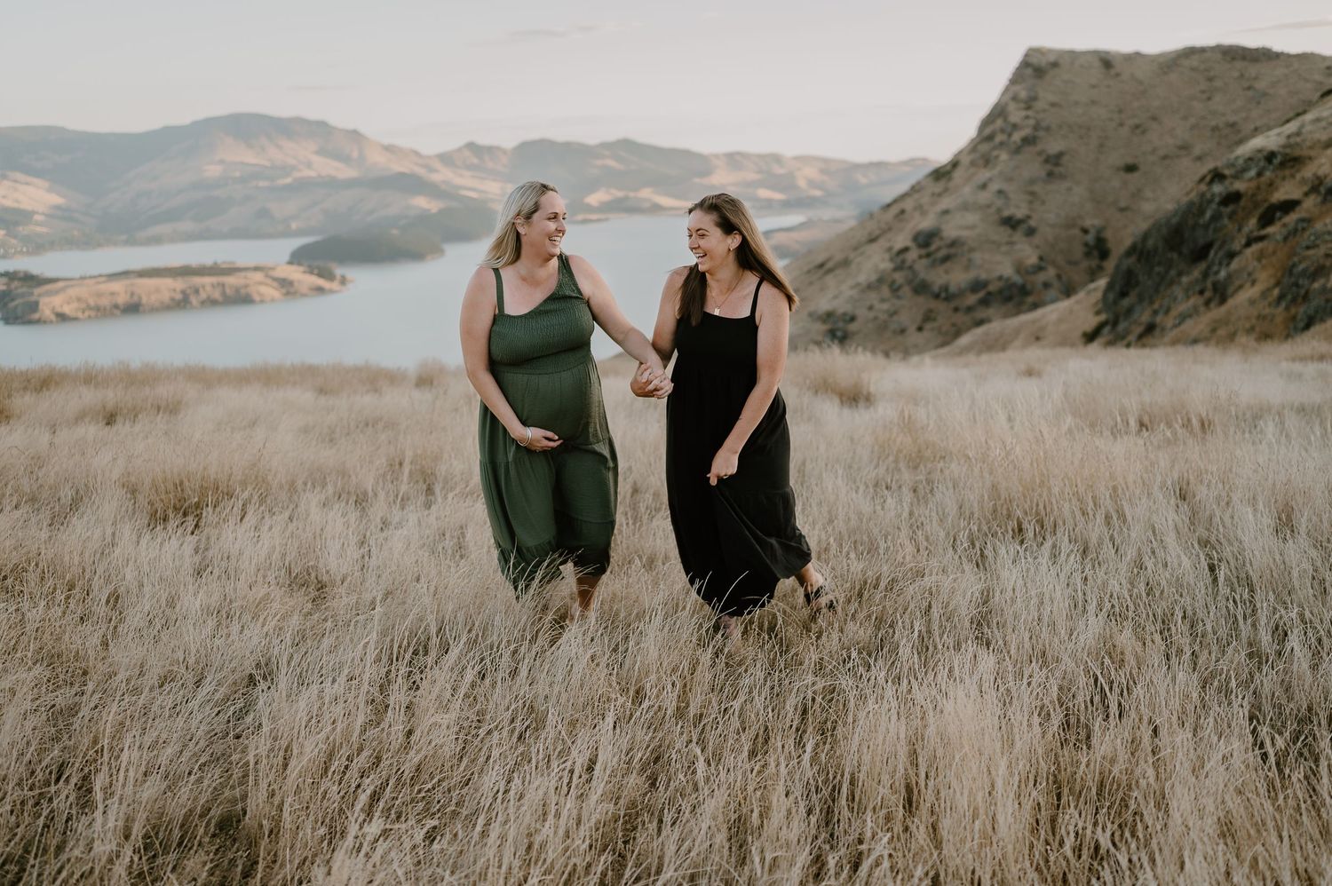 Two women in flowing dresses walking through golden grass with mountain lake backdrop.