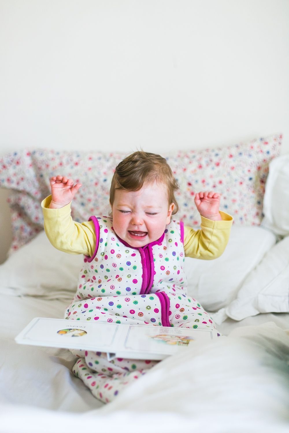 A happy baby in yellow and pink polka dot pajamas expressing joy on a white bed.