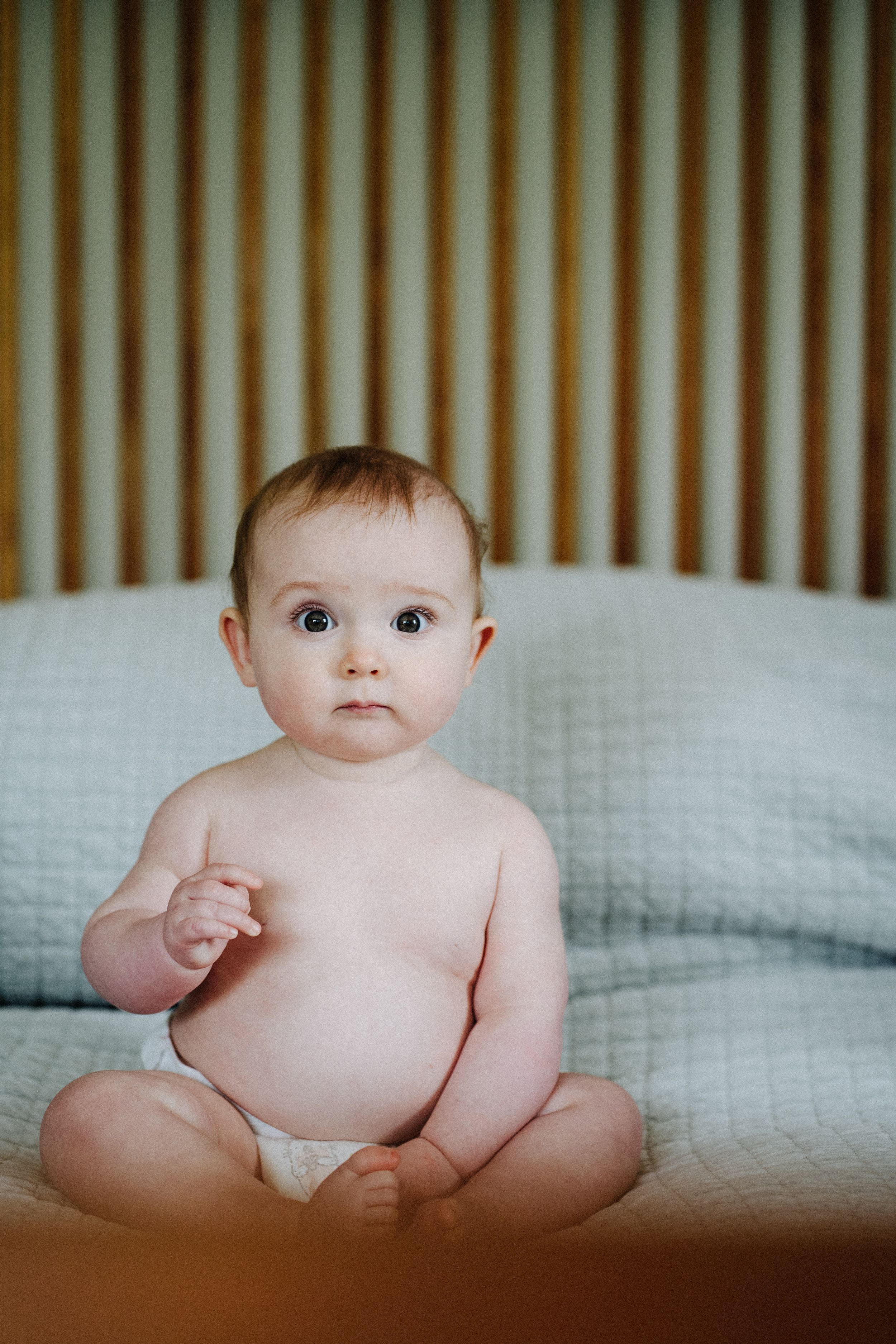 A baby in white diaper sits upright on textured bedding against a wooden headboard.