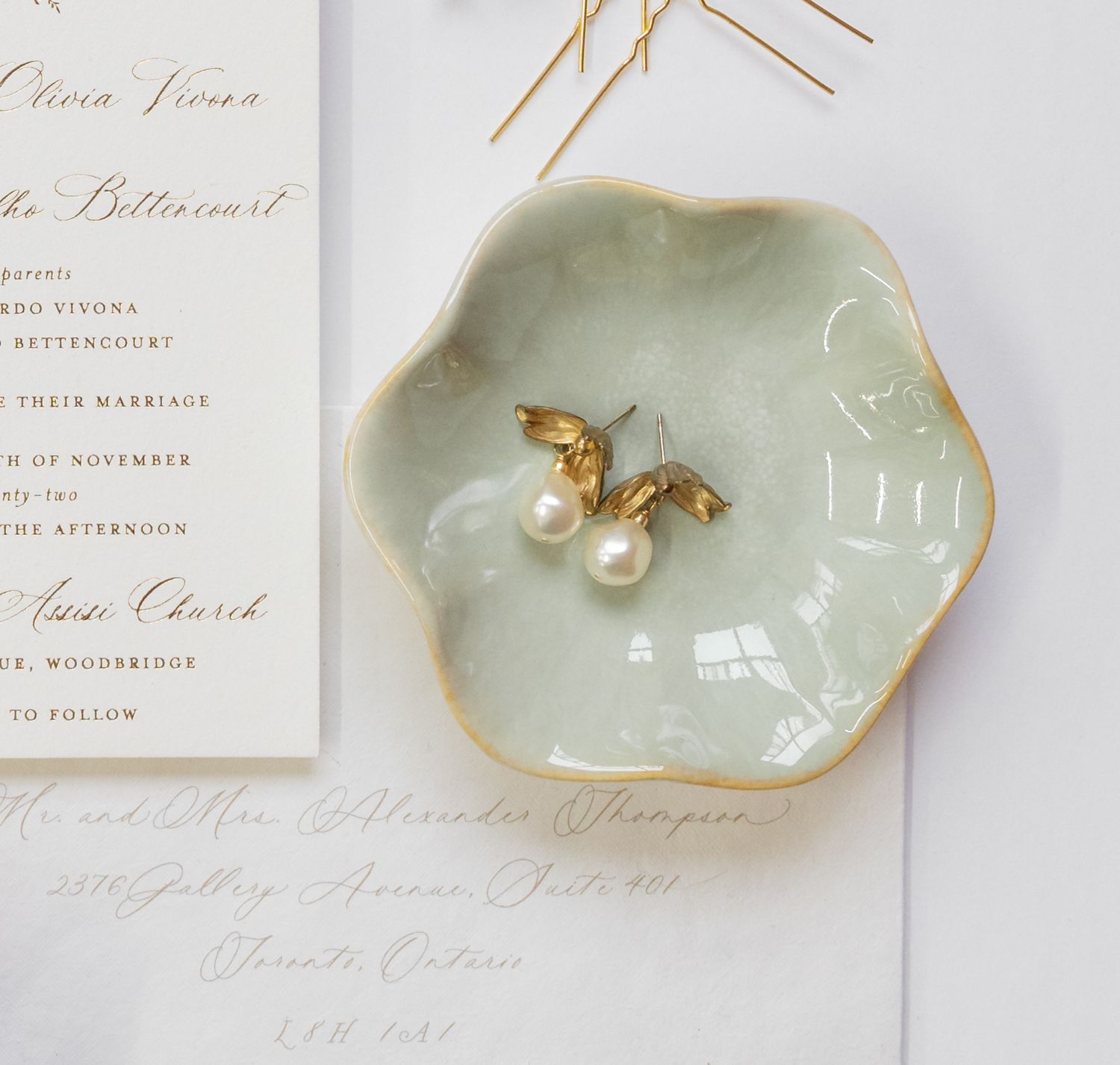 Gold pearl earrings displayed on mint ceramic trinket dish next to wedding invitation.