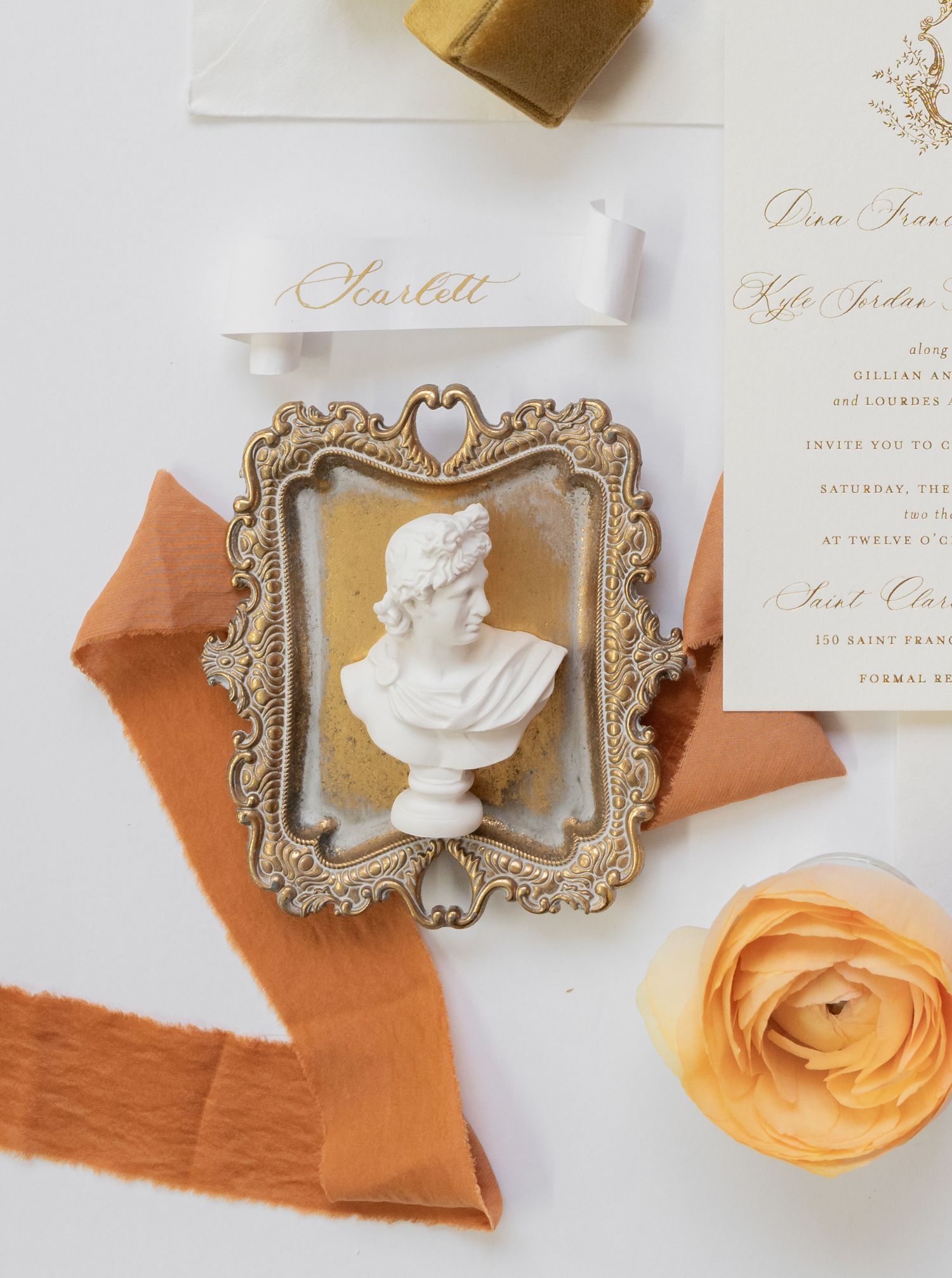 Wedding stationery with ornate gold frame, antique bust sculpture, and orange silk ribbon.