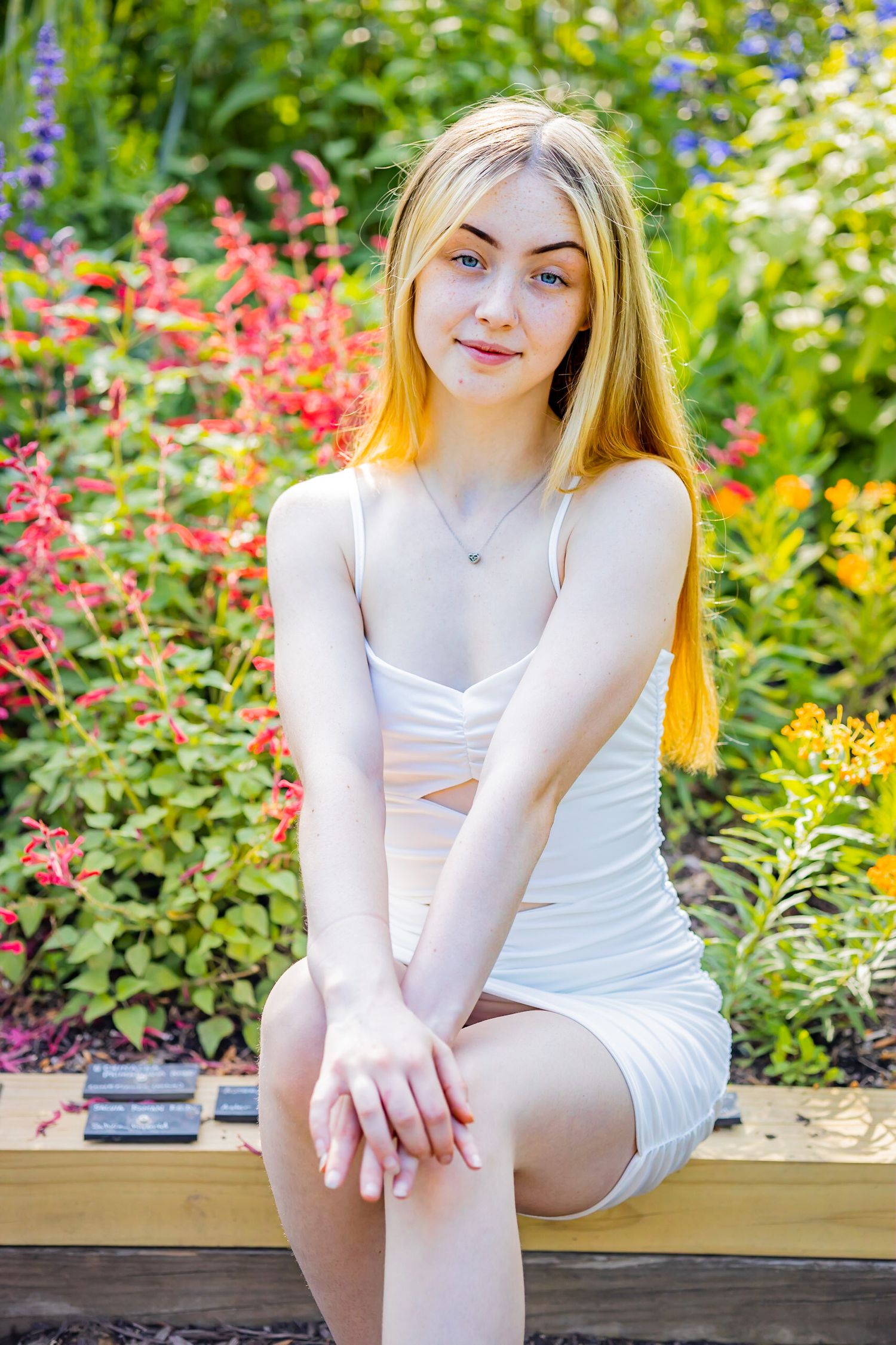 A graduate in white dress sitting on bench surrounded by colorful garden flowers.
