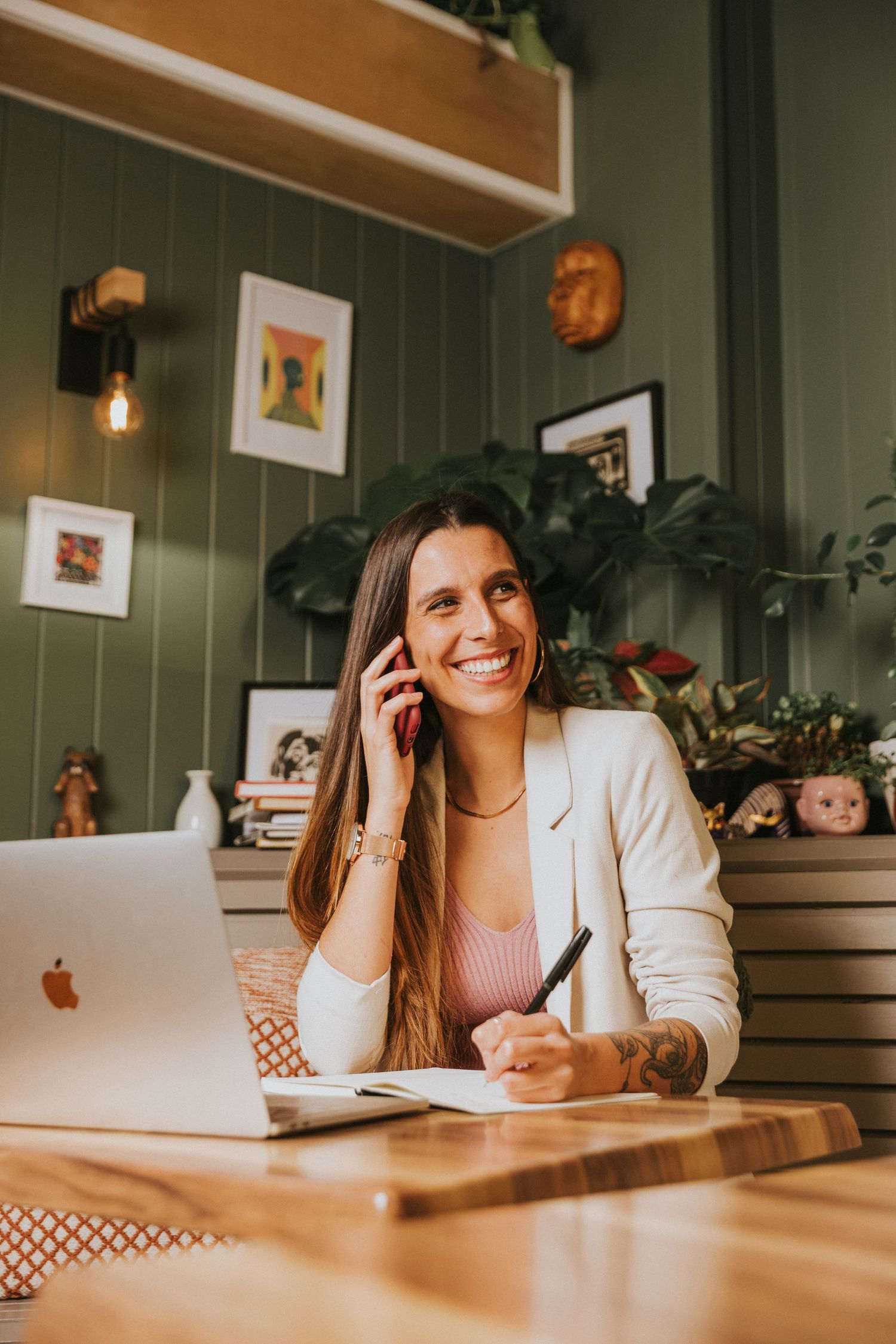 Woman working remotely at desk with laptop surrounded by plants and artwork in cozy home office setting.