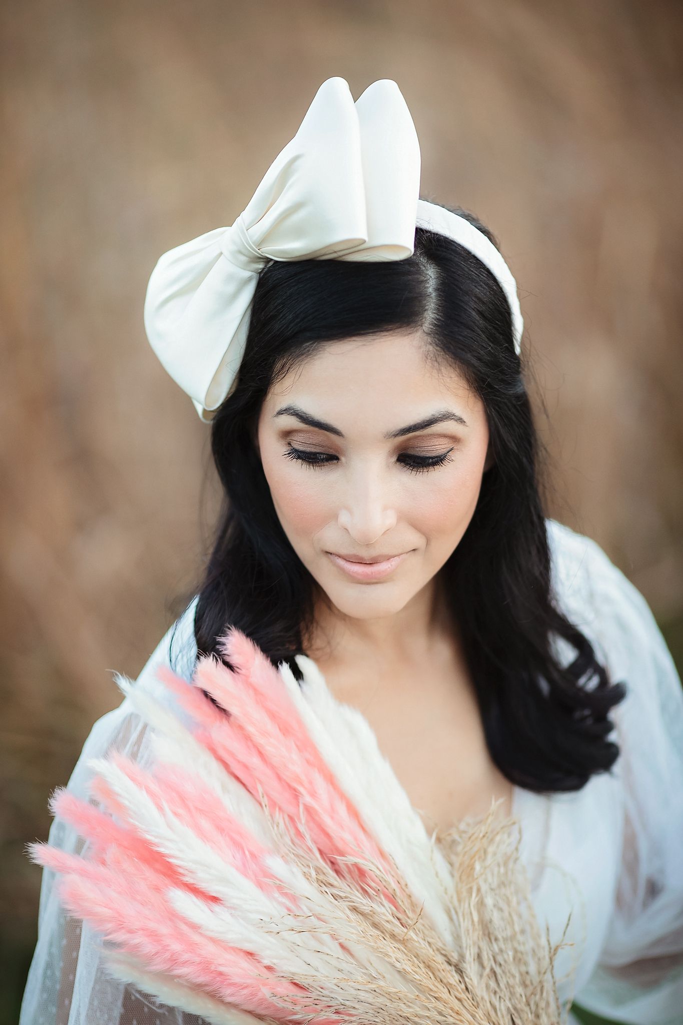 Bride with dark hair wearing a white satin bow headband, holding pink pampas grass bouquet outdoors.