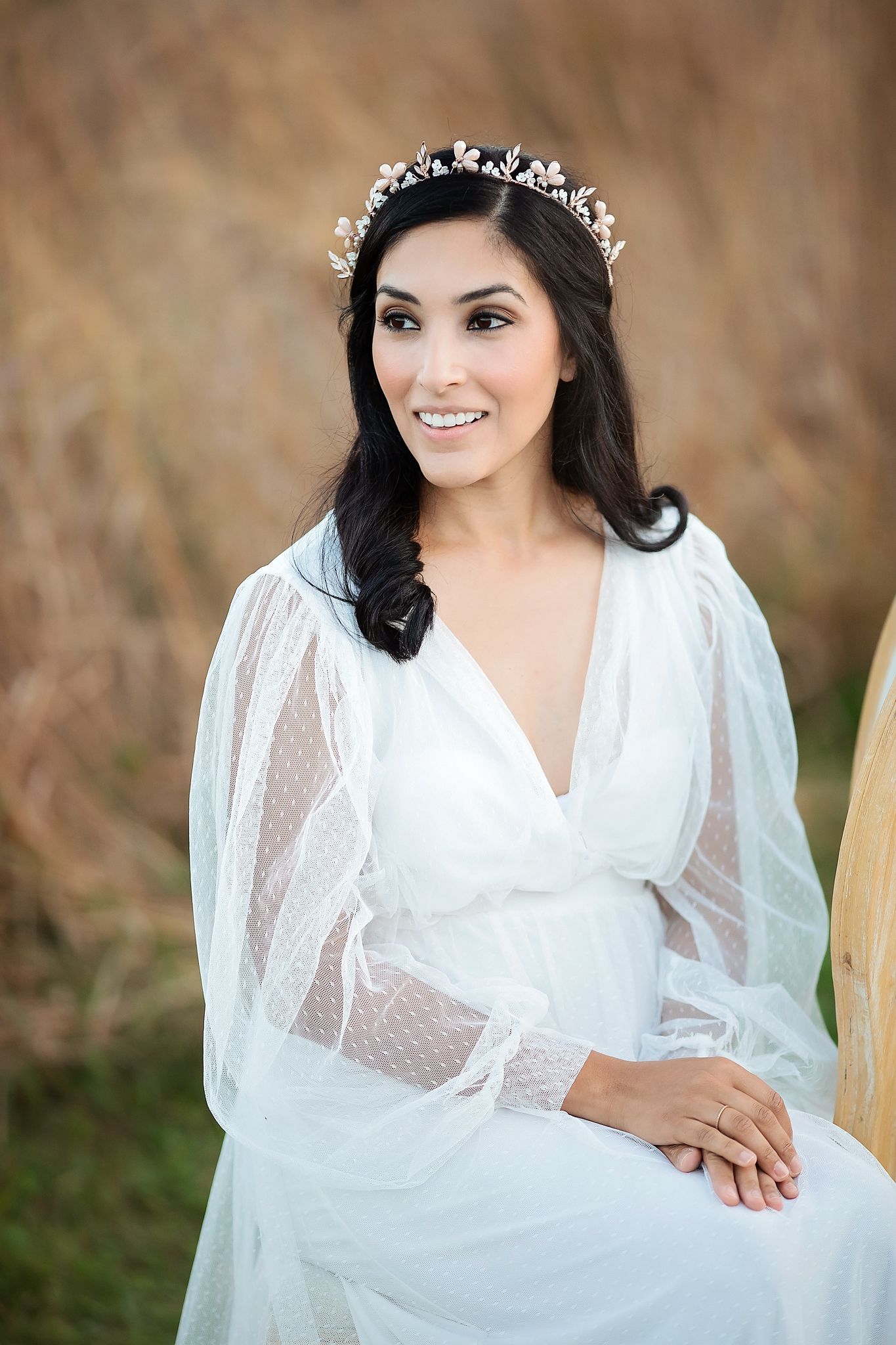 Elegant bride in sheer white dress wearing delicate floral crown tiara, seated outdoors in field.