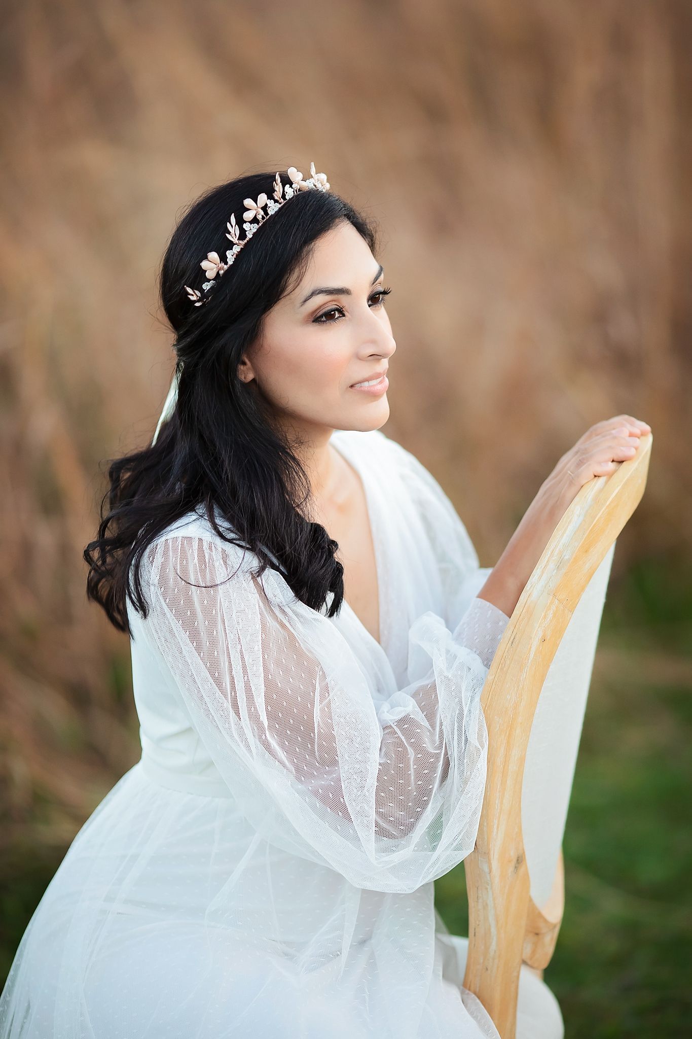 Bride in sheer white gown with gold floral tiara holding vintage chair in golden grass field.