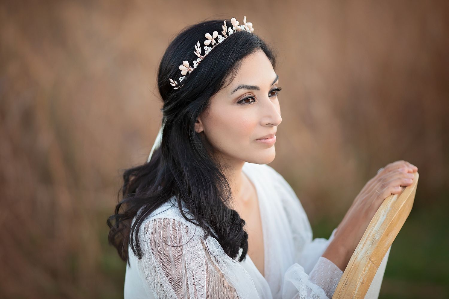 Dark-haired bride wearing ornate gold floral crown tiara, posing with vintage chair in field.