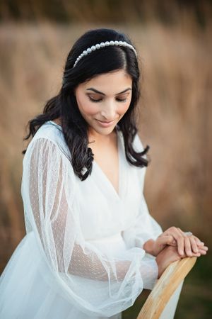Elegant bride in sheer white dress wearing pearl headband, looking down while leaning on chair.