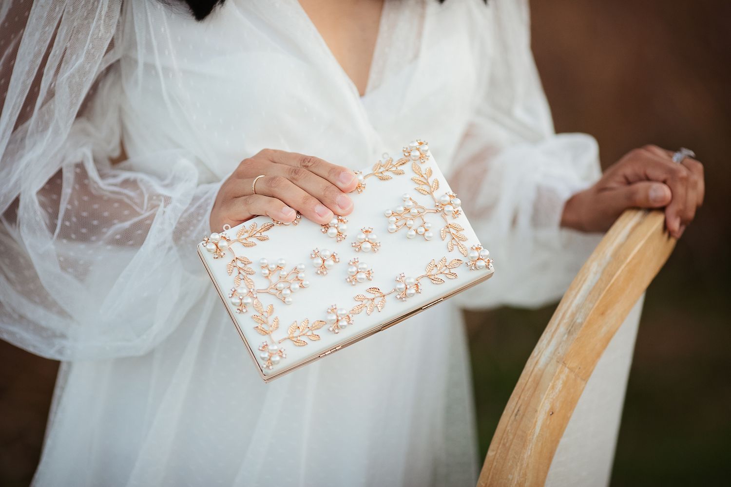 Close-up of a bride holding an ornate white and gold floral embroidered clutch purse in her wedding dress.