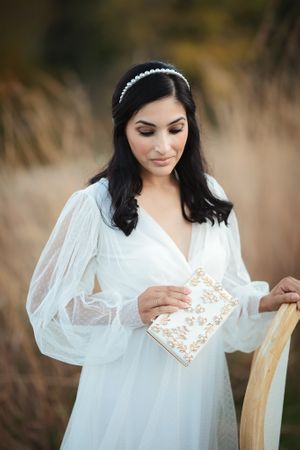 Elegant bride in white sheer dress holding a gold floral clutch while standing in a golden field.