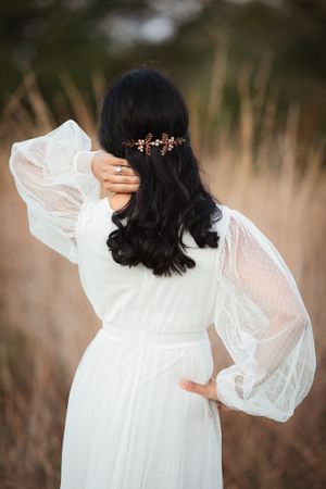 Back view of a bride in white sheer dress touching her rose gold floral hair comb in a field.