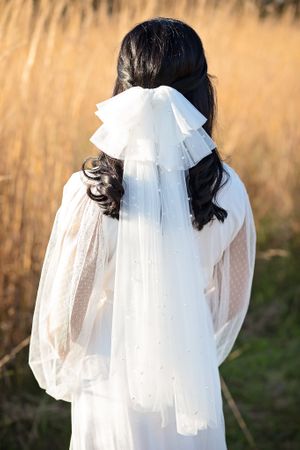 Back view of a bride in white dotted sheer dress with a pearl tulle bow veil in a sunlit field.