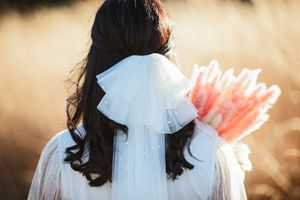 Back view of a bride with a white pearl tulle bow veil holding pink feathers in a golden field.