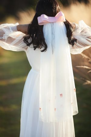 Back view of a bride in a white dress with a pink bow veil detail in a sunlit outdoor setting.
