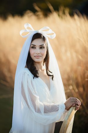Elegant bride in a white lace wedding dress with a white bow veil posing in golden wheat fields.