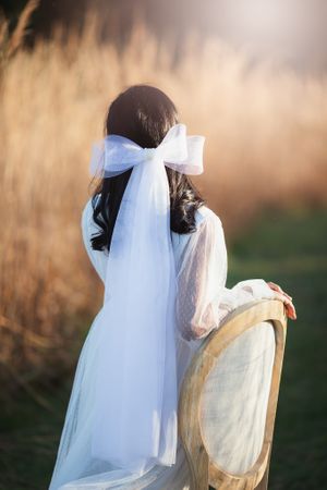 Back view of a bride in a white wedding dress with a white bow veil beside a vintage chair outdoors.