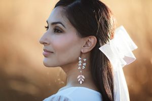 Side profile of a bride wearing crystal leaf drop earrings and a white bow veil in golden light.