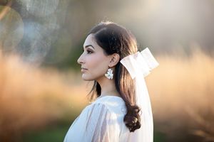 Elegant bride in profile wearing a white bow veil and floral earrings in a sun-drenched field.