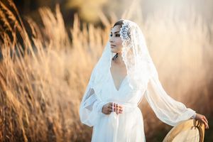 Bride in white dress and lace veil standing in golden wheat field at sunset.