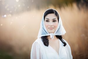 Portrait of dark-haired bride in white lace hood veil in sunny field.
