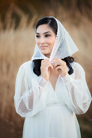 Smiling bride adjusting white pearl-trimmed mantilla veil in golden field.