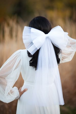 Back view of a dark-haired bride wearing a large white tulle bow veil and polka dot lace dress outdoors.