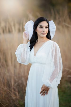 Elegant woman in a white dotted lace dress with sheer sleeves holding a ruffled bonnet in golden field.