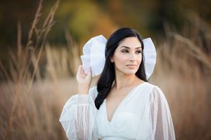Beautiful dark-haired woman in white lace dress holding a ruffled bonnet in a golden wheat field.