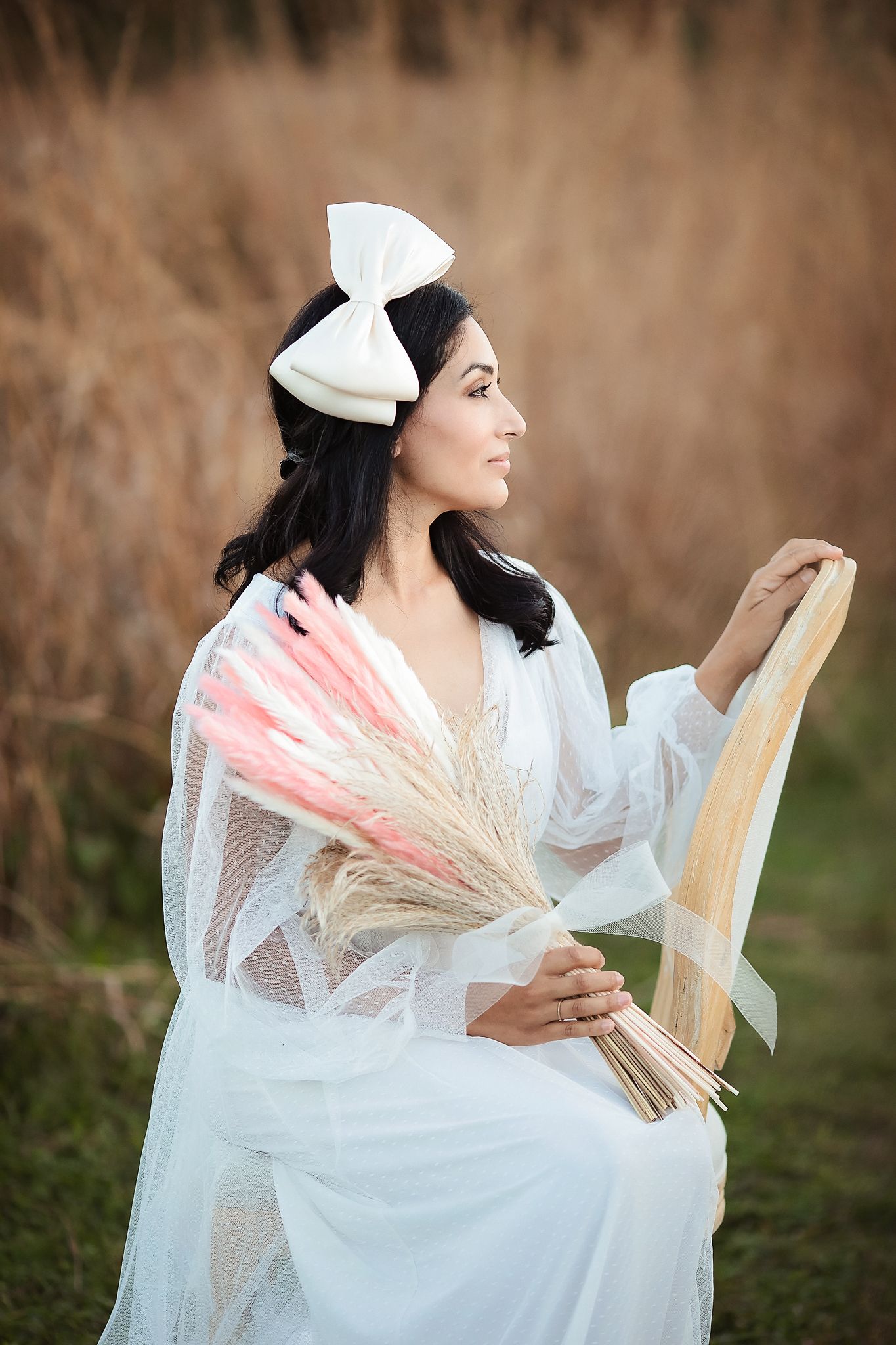 Serene bride in white lace dress with bow headband holding dried floral bouquet while seated in a field.