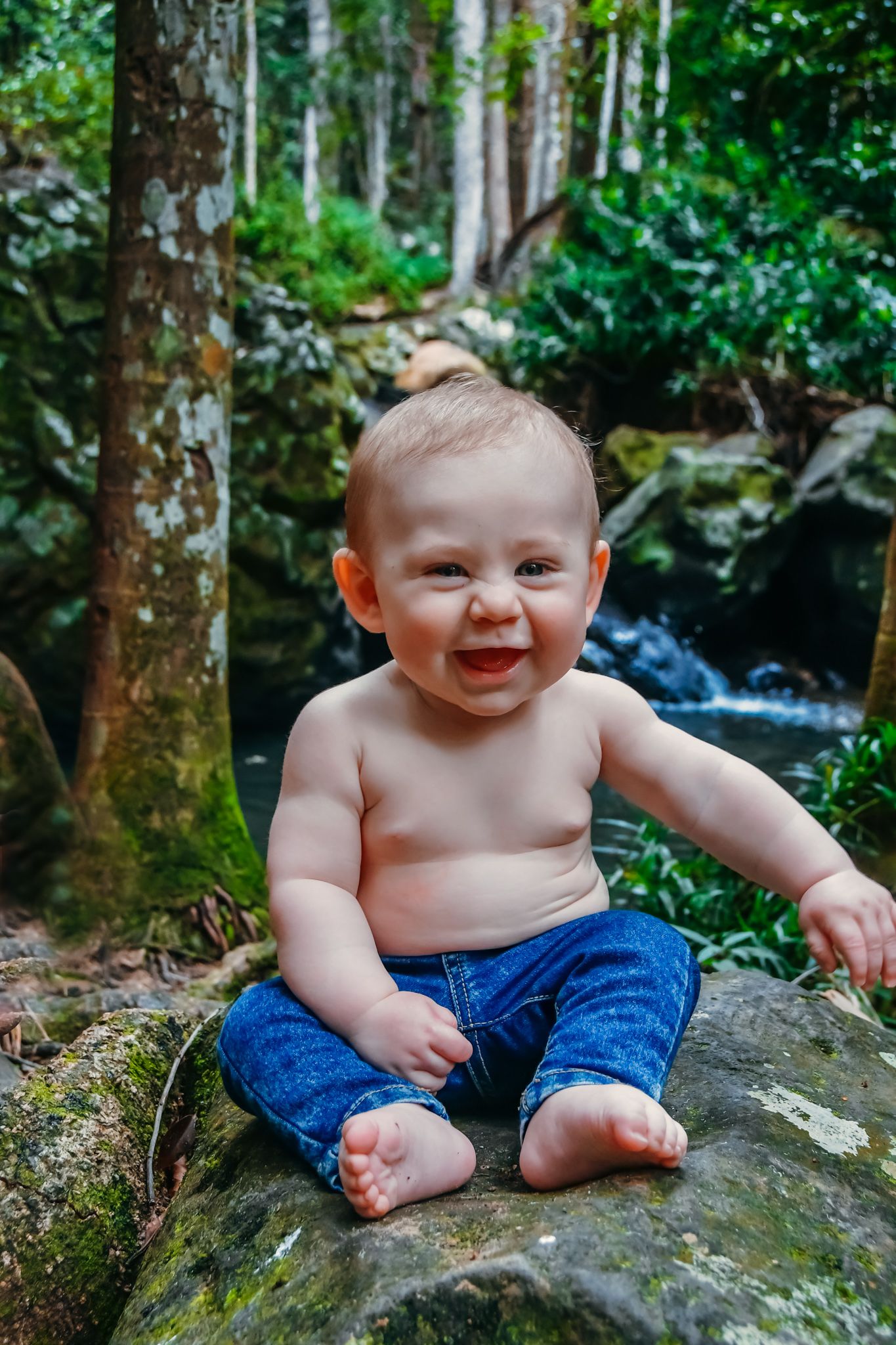 Baby wearing blue shorts sits on mossy rocks near a forest waterfall.