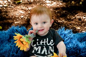 Young child in 'Little Man' shirt holds sunflower while sitting on bright blue blanket.