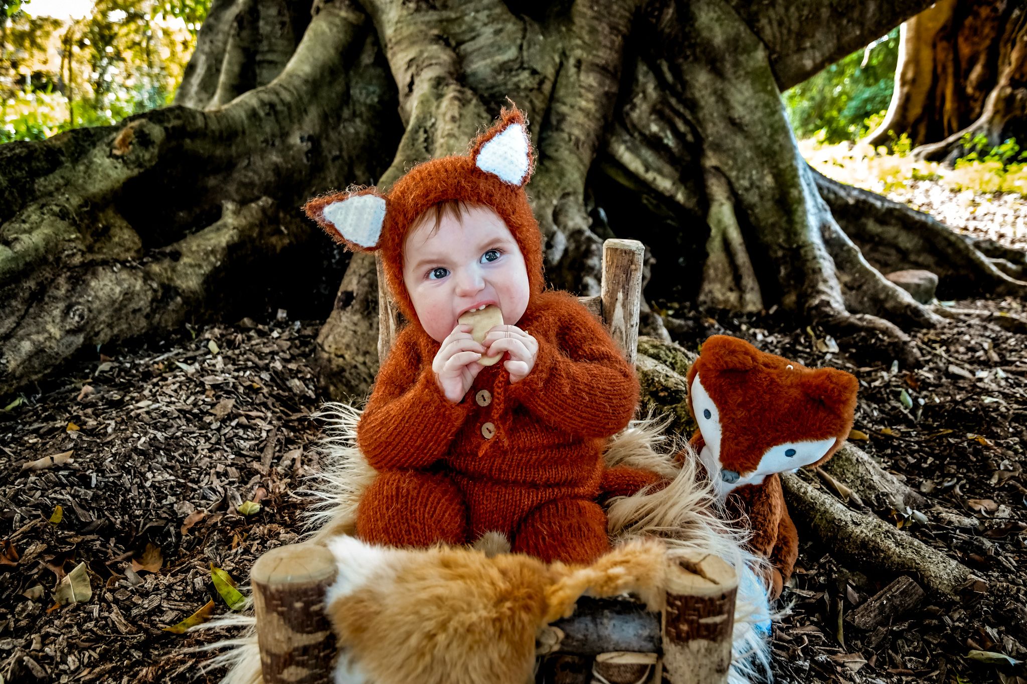 Small figure in orange sweater sits under large tree roots on furry blanket with woodland decorations.