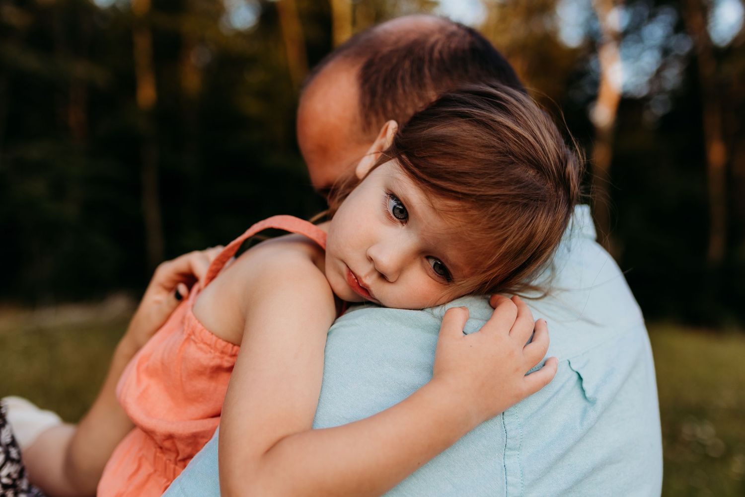 Child resting head on shoulder in warm evening sunlight through trees.