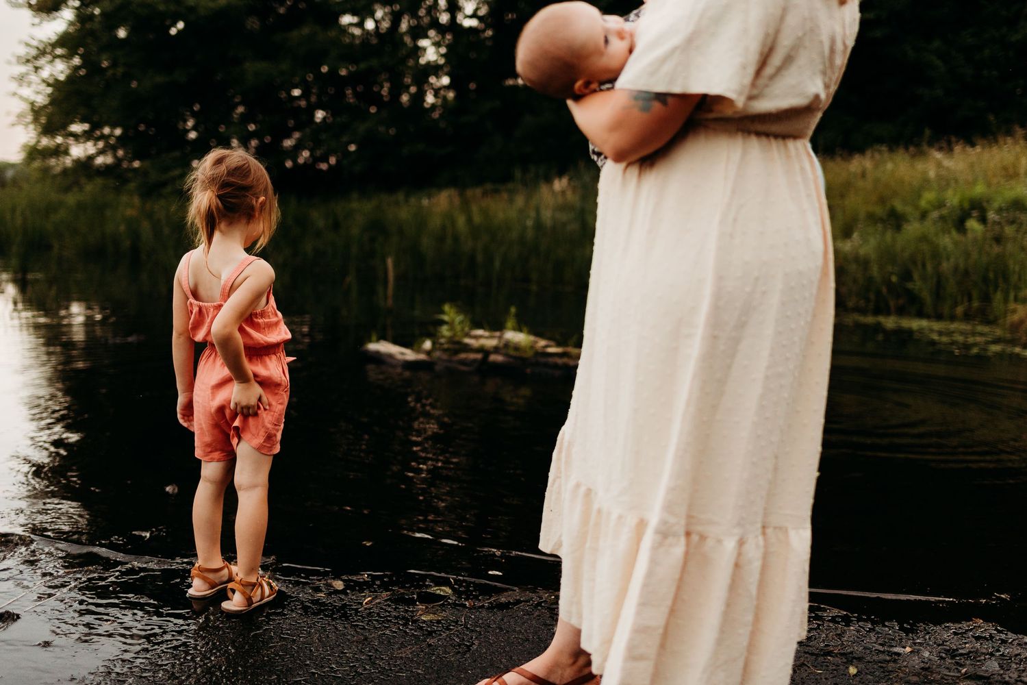 Two people standing by a serene lake edge in summer dresses during sunset.