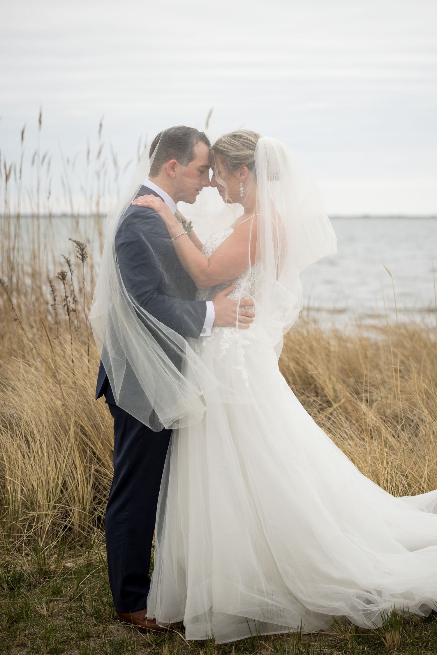A romantic wedding couple embraces in tall beach grass with flowing veil and ocean backdrop.
