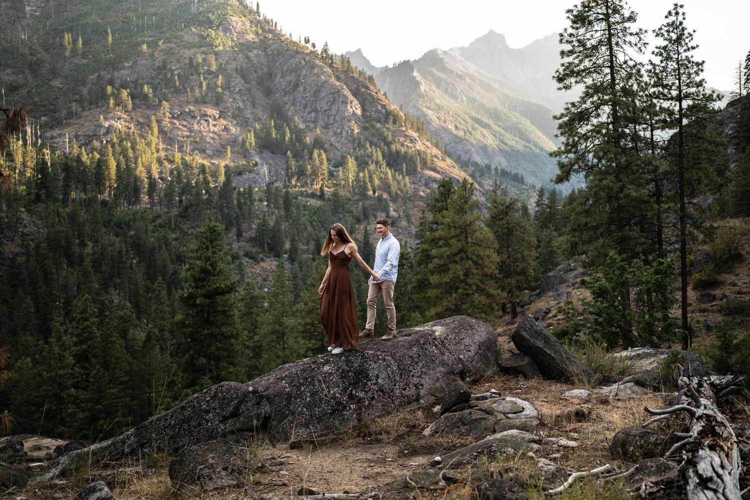 Couple standing on rock overlook with scenic mountain valley and evergreen forest backdrop.