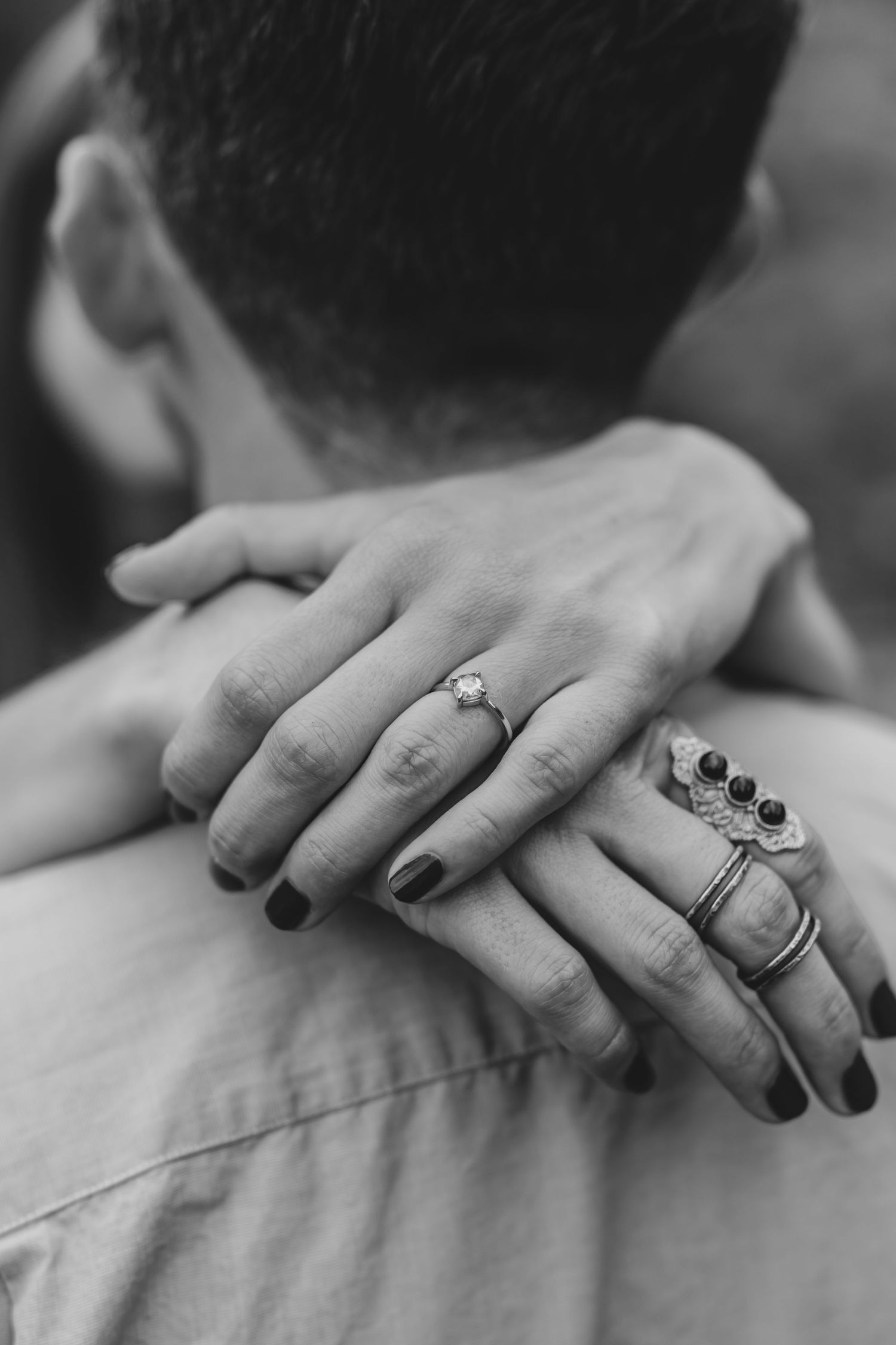 Close-up black and white photo of hands with rings embracing neck showing intimate connection and jewelry details.