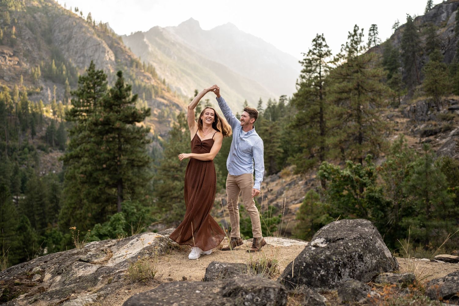 Dancing couple on mountain ledge surrounded by evergreen forest and majestic peaks at sunset.