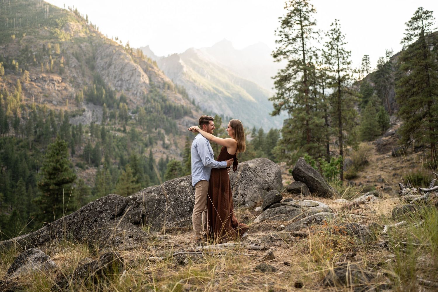 Romantic couple embracing on mountain cliff with stunning mountain landscape and pine trees in background.