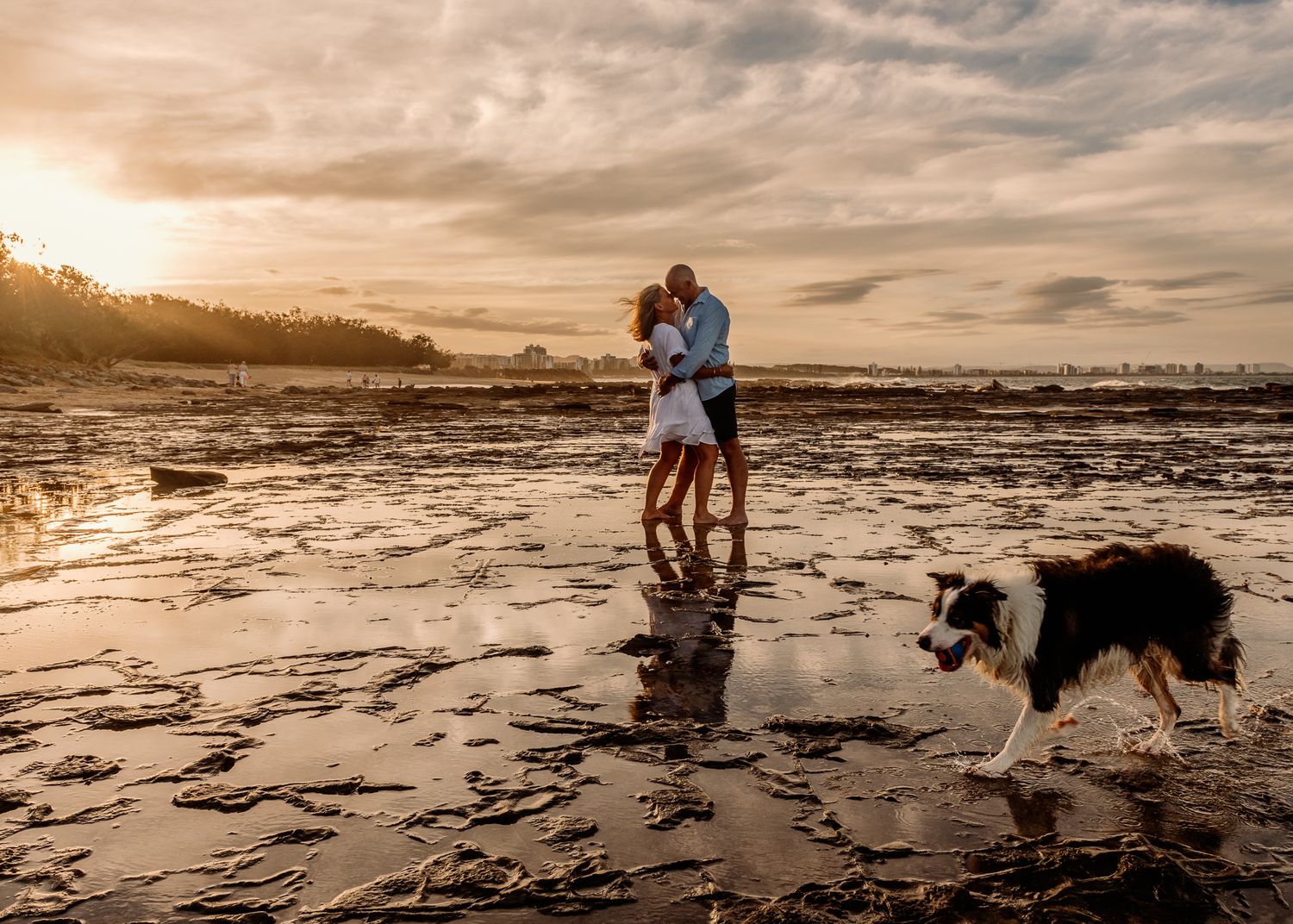 A couple embraces on a beach at sunset while their Border Collie walks nearby.