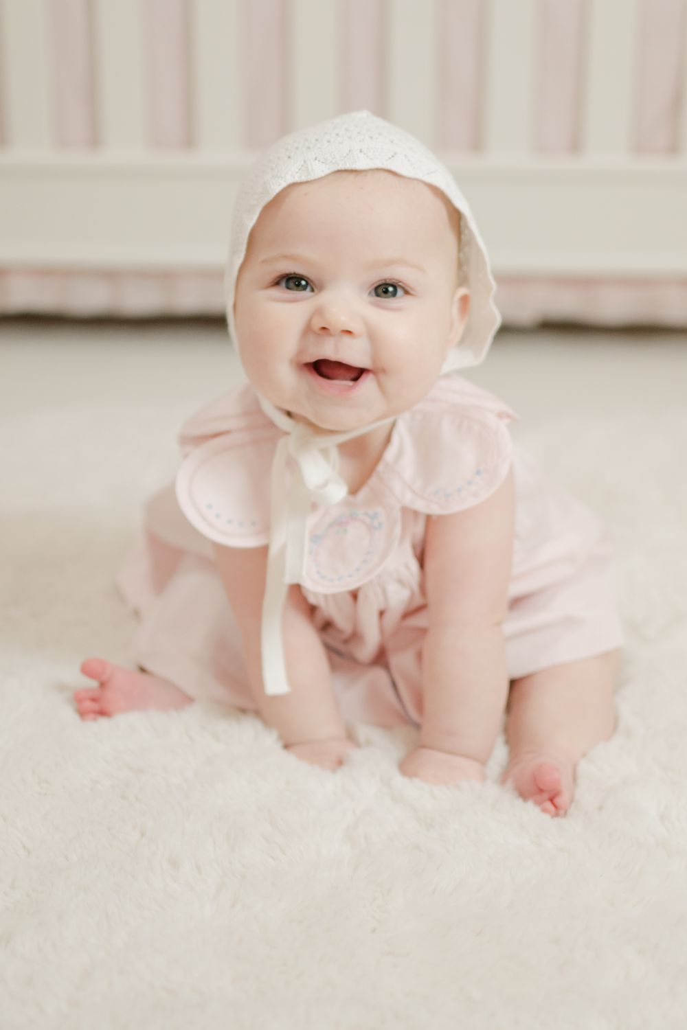 Happy baby girl in pink outfit and white bonnet sitting up and smiling on a fluffy white rug.