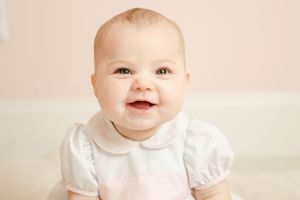 Smiling baby with green eyes wearing a white peter pan collar outfit against a soft pink background.