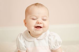 Laughing baby with eyes closed wearing a white peter pan collar outfit against a soft pink background.