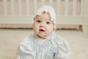 Cheerful baby girl in blue smocked dress and white lace bonnet sitting and smiling at camera.