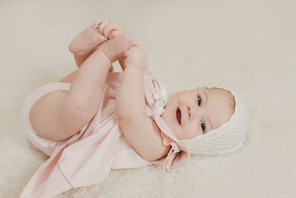 Adorable baby girl in pink dress and white knit bonnet lying on fluffy white rug holding her feet.