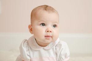 Close-up portrait of a baby in a white and pink peter pan collar outfit against a soft background.