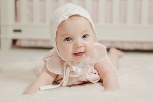Cute baby girl in pink dress and white bonnet doing tummy time on a soft white fluffy rug.