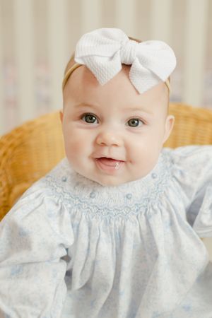 Smiling baby girl in blue smocked dress with white bow headband sitting in a wicker chair.